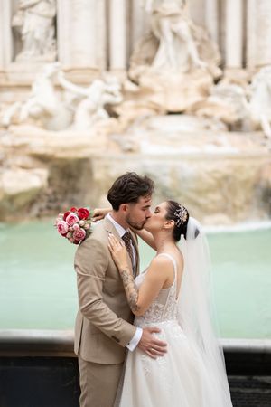 A romantic wedding couple shares a kiss in front of the historic Trevi Fountain in Rome.