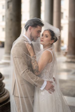 A romantic wedding portrait of a couple embracing under elegant columns, with the bride wearing a flowing veil and lace gown.