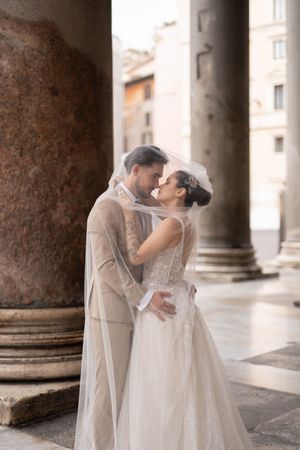 A newlywed couple embraces under classical stone columns in an elegant European setting.