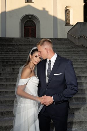 A couple in wedding attire share an intimate moment on stone steps outside a church.