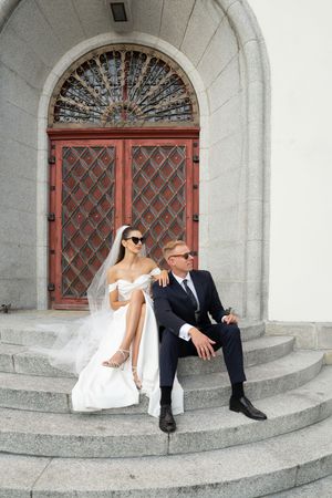 A couple in wedding attire sits casually on stone church steps in a series of candid poses.