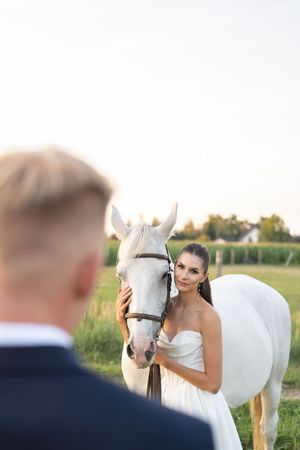 A bride in a white dress shares a tender moment with a white horse in a sunny meadow at sunset.