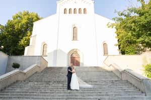 A white church with stone steps leading up to its entrance sits nestled among green trees on a sunny day.