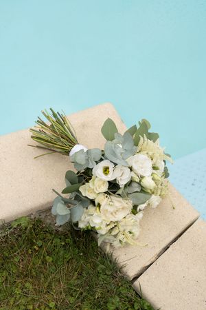A white floral bouquet with eucalyptus leaves rests on a concrete surface against a bright blue sky background.