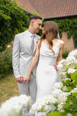A couple in wedding attire share an intimate moment among white hydrangea bushes beside a brick house.