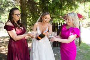 Bride in white gown pours champagne with two bridesmaids in pink and burgundy dresses outdoors.