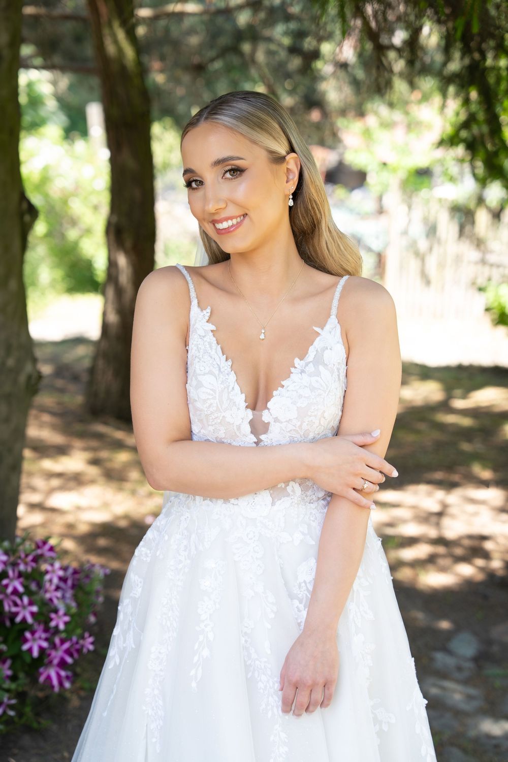 Smiling bride in a white lace V-neck wedding gown with floral appliques, posing outdoors among trees and pink flowers.