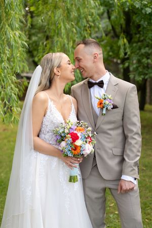 A bride and groom smile at each other outdoors, she holds a colorful bouquet, he wears a tan suit with bow tie.