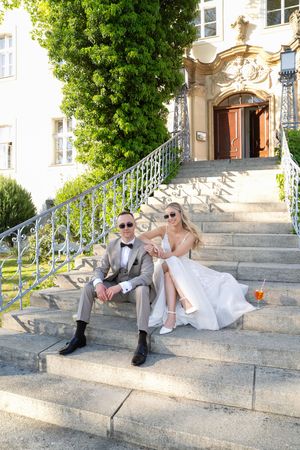 Bride and groom sitting on elegant stone steps outside a classic building, wearing sunglasses and holding champagne.