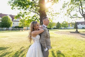 Bride in white lace gown embraces groom in tan suit outdoors during golden hour wedding portrait.