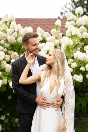 A romantic couple embraces near blooming white hydrangea bushes against a brick wall backdrop.