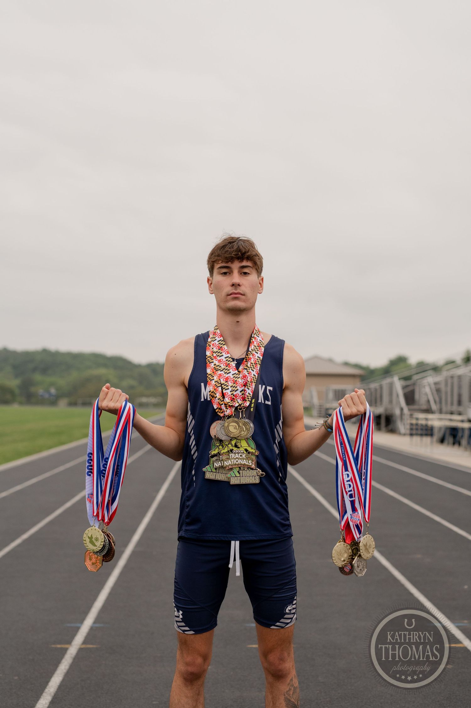 Track athlete poses with multiple medals and ribbons on outdoor running track.