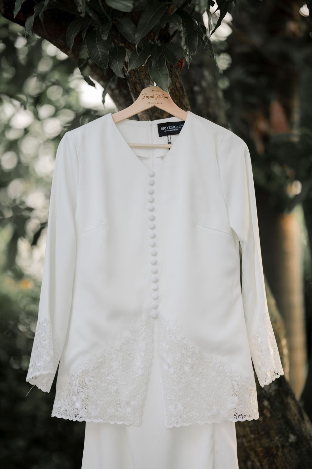 Elegant white wedding dress jacket with lace trim and button details hanging on wooden hanger against natural foliage.