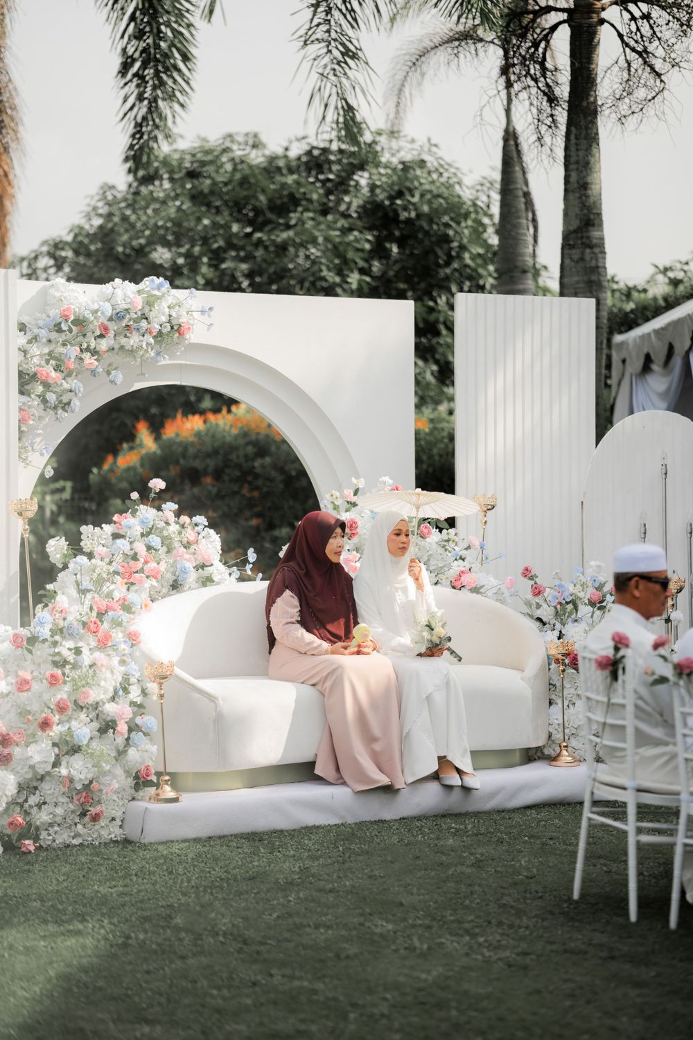 Elegant white floral arch setup with couch for outdoor wedding ceremony against palm tree backdrop.