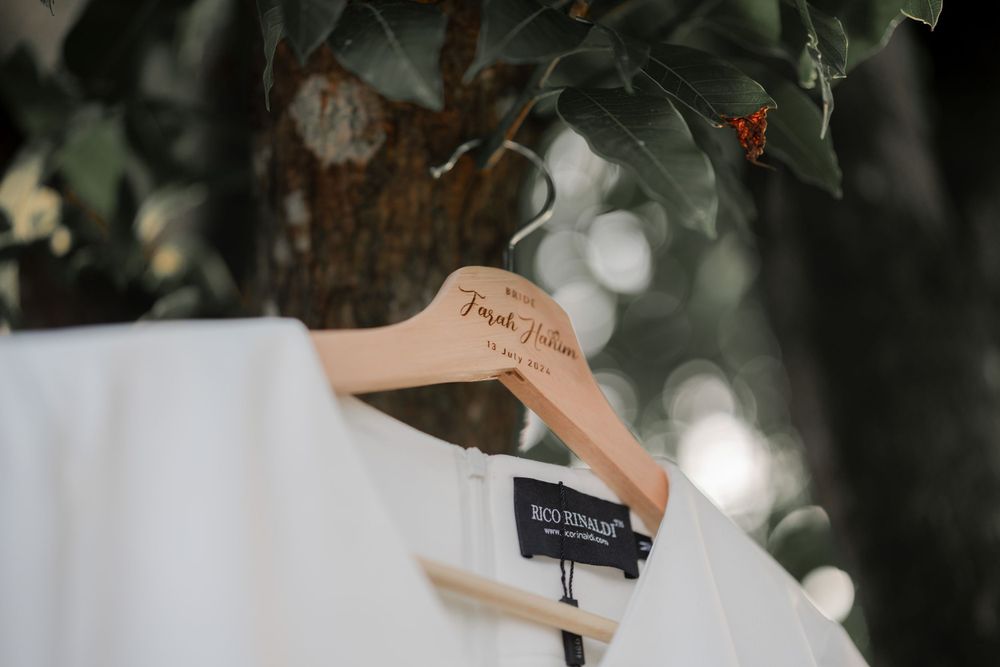 A white wedding dress hangs from a wooden hanger against a bokeh nature background.