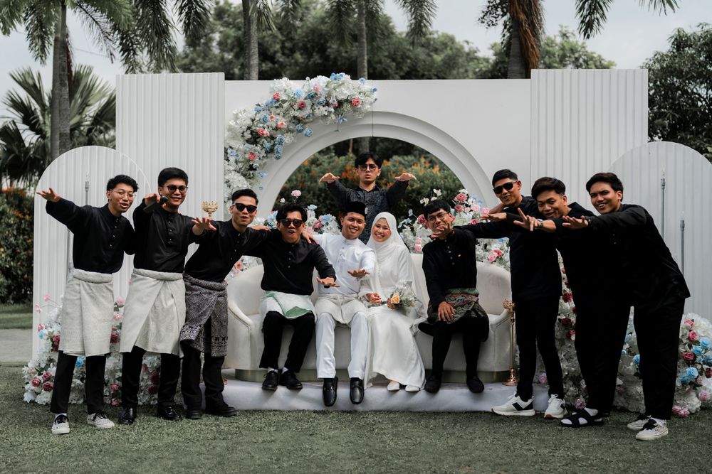 Wedding party group photo under white floral arch in outdoor garden setting.