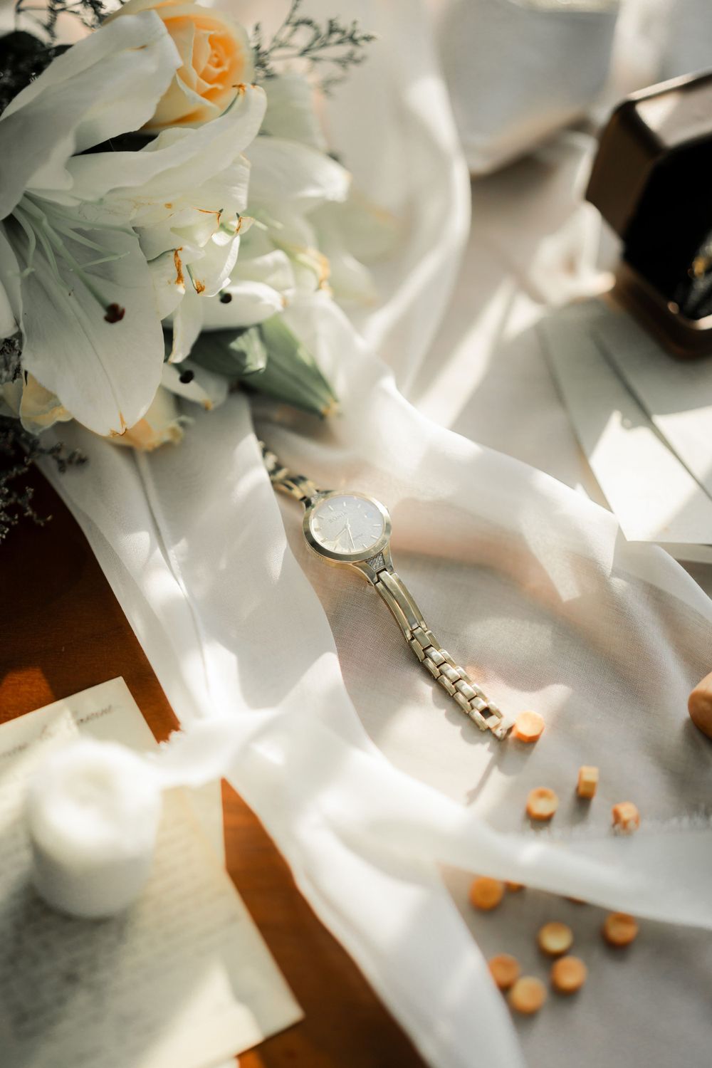 Wedding details flatlay with white lilies, engagement ring, and scattered pearls on white silk fabric.