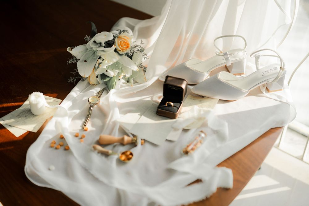 Bridal accessories including jewelry box, shoes, and floral bouquet arranged on white fabric in sunlight.