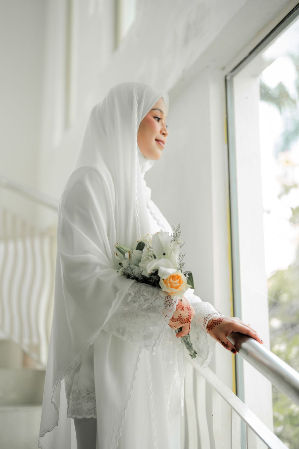 Bride in elegant white hijab and dress stands by window holding delicate peach and white floral bouquet.