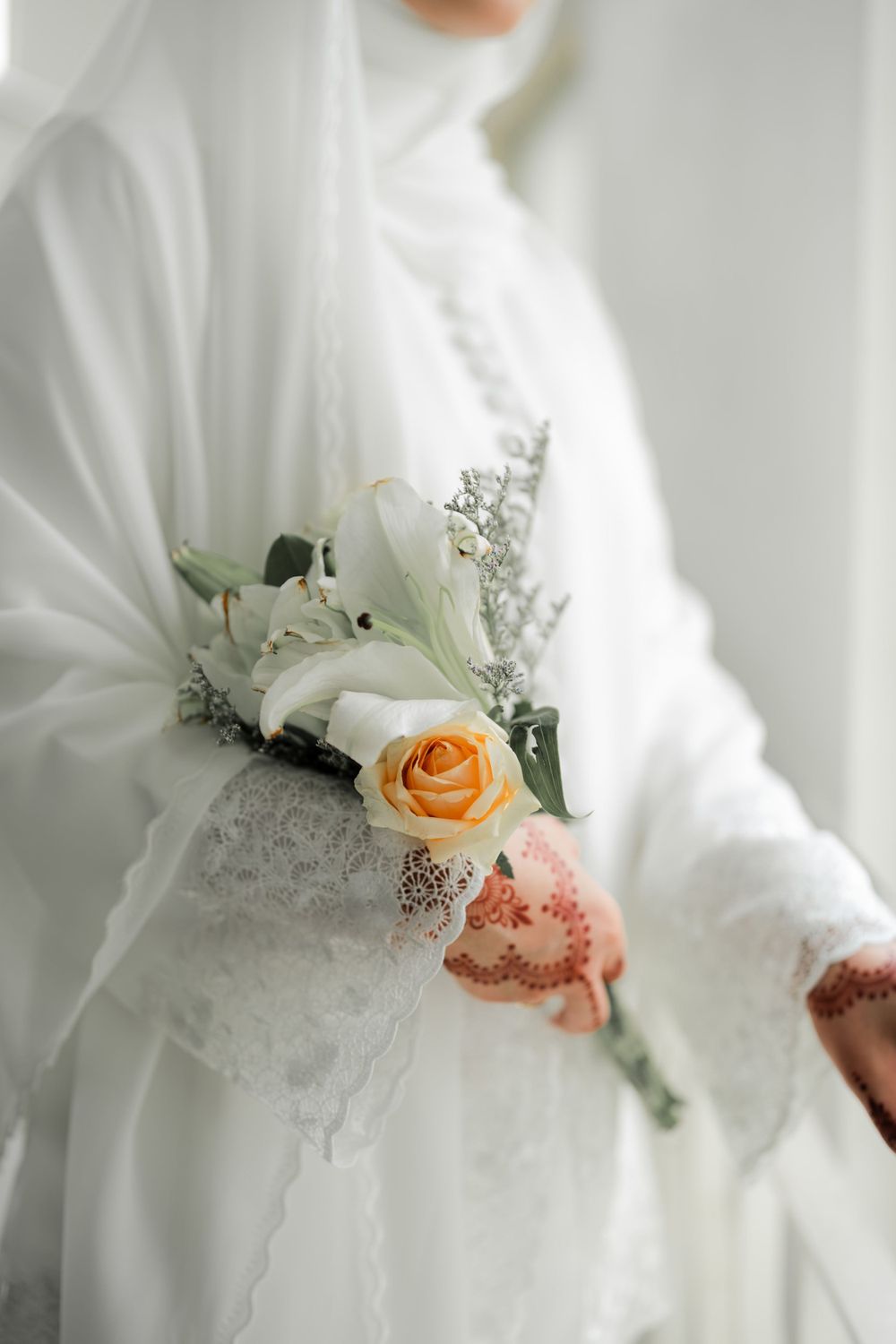 Close-up of a delicate peach rose and white lily bouquet against white wedding dress with lace details.