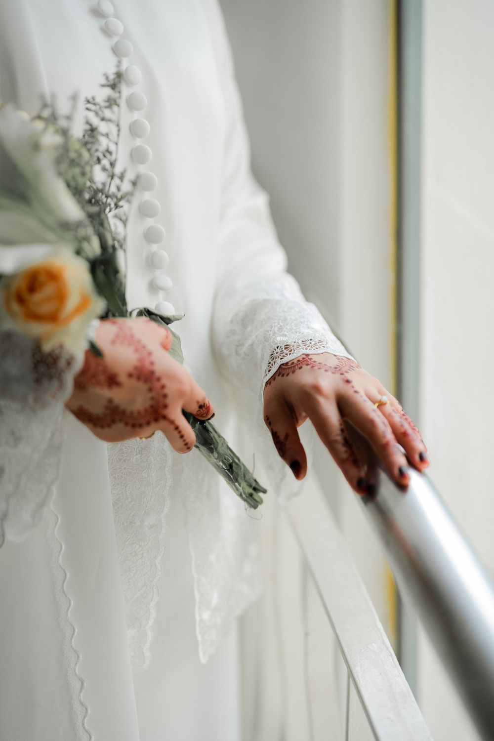 Close-up of henna-adorned hands holding onto a white railing, with wedding dress and floral details visible.