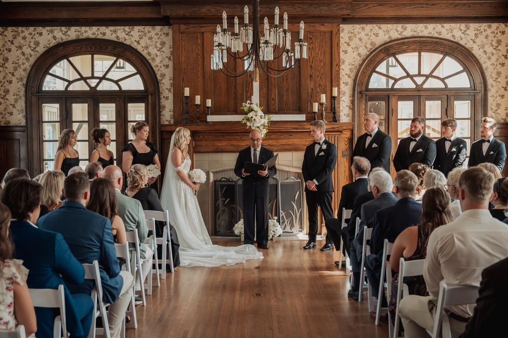 A panoramic view of a wedding ceremony in an elegant wood-paneled ballroom with arched windows and chandeliers.