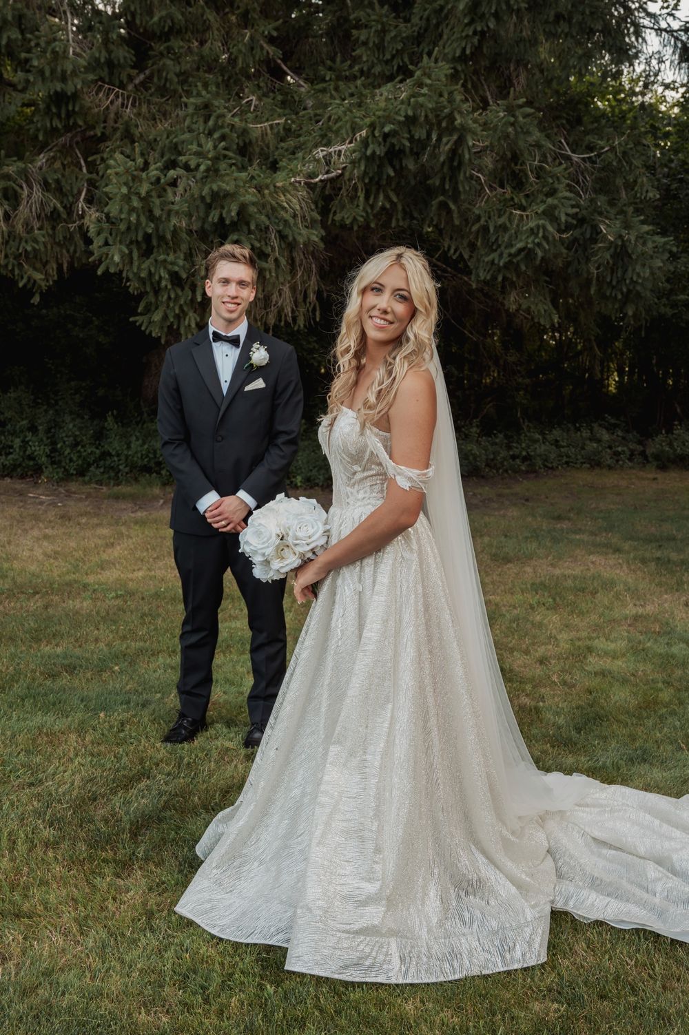A wedding couple poses together outdoors on a grassy lawn with trees in the background, the bride wearing a flowing white gown.