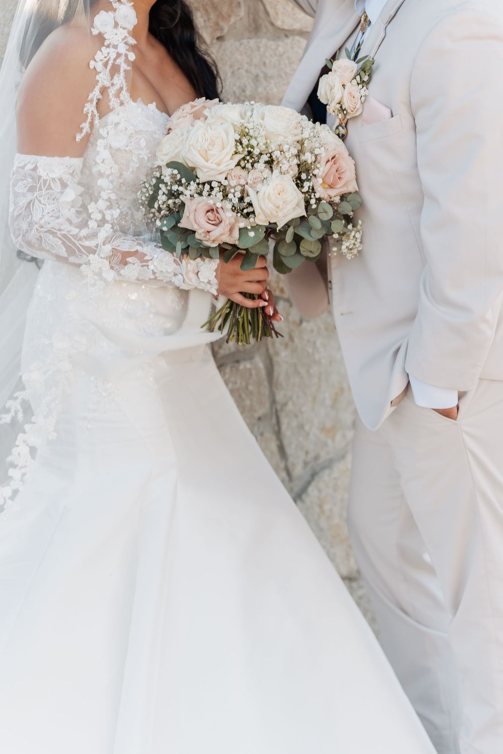 Elegant wedding couple holds white and blush pink floral bouquets against a rustic stone wall backdrop.