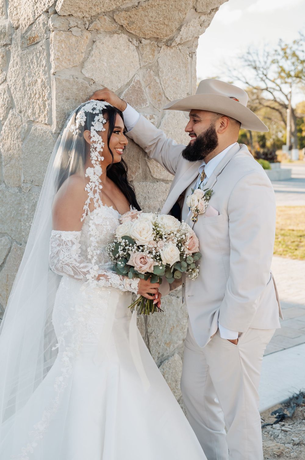 A romantic wedding photo showing a couple in light-colored attire posing against a stone wall with a beautiful bouquet.