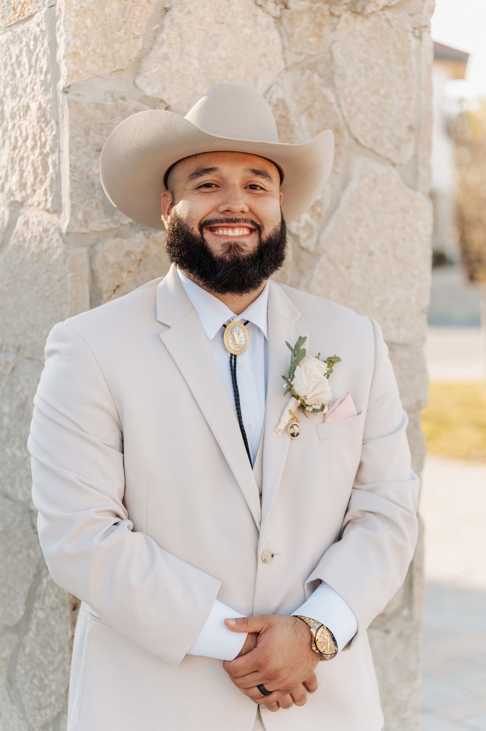 A groom in a light beige suit and cowboy hat smiles while wearing a floral boutonniere and bolo tie at an outdoor venue.