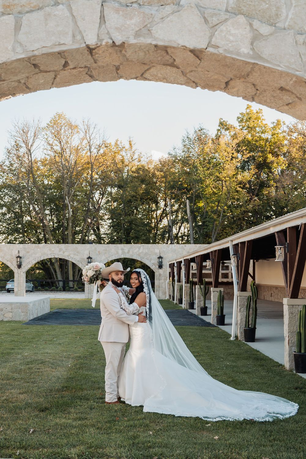 A wedding couple shares romantic moments under a white tent canopy in an outdoor garden setting at sunset.