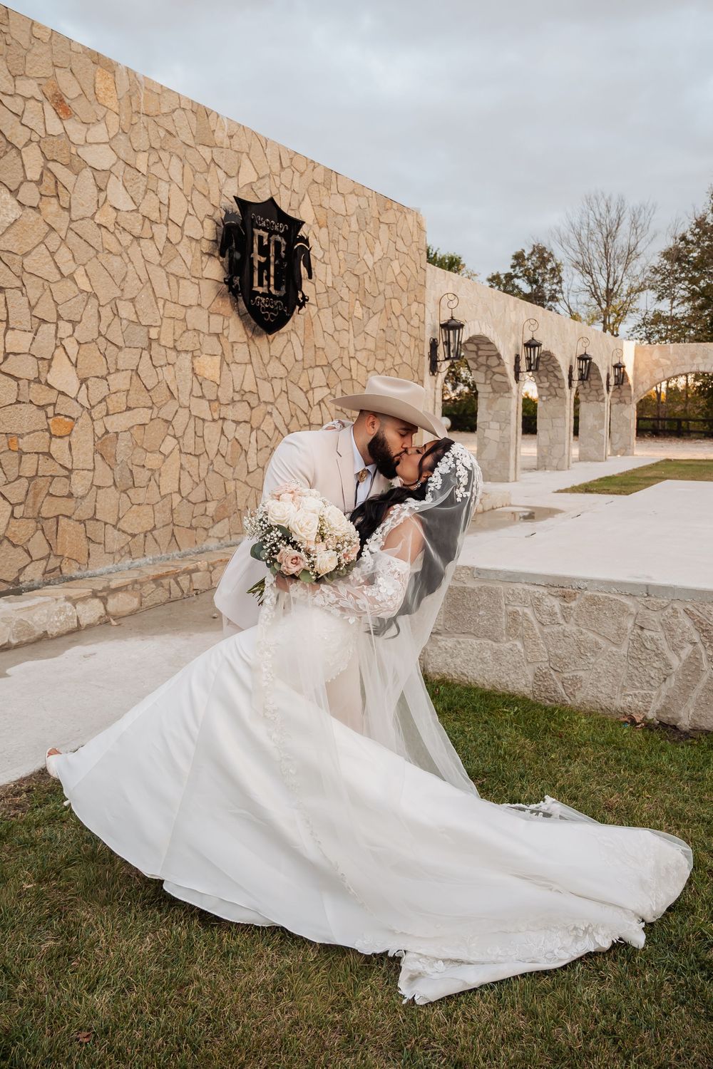 A romantic wedding moment captured against a stone wall with arches, featuring a flowing white dress and cowboy hat.