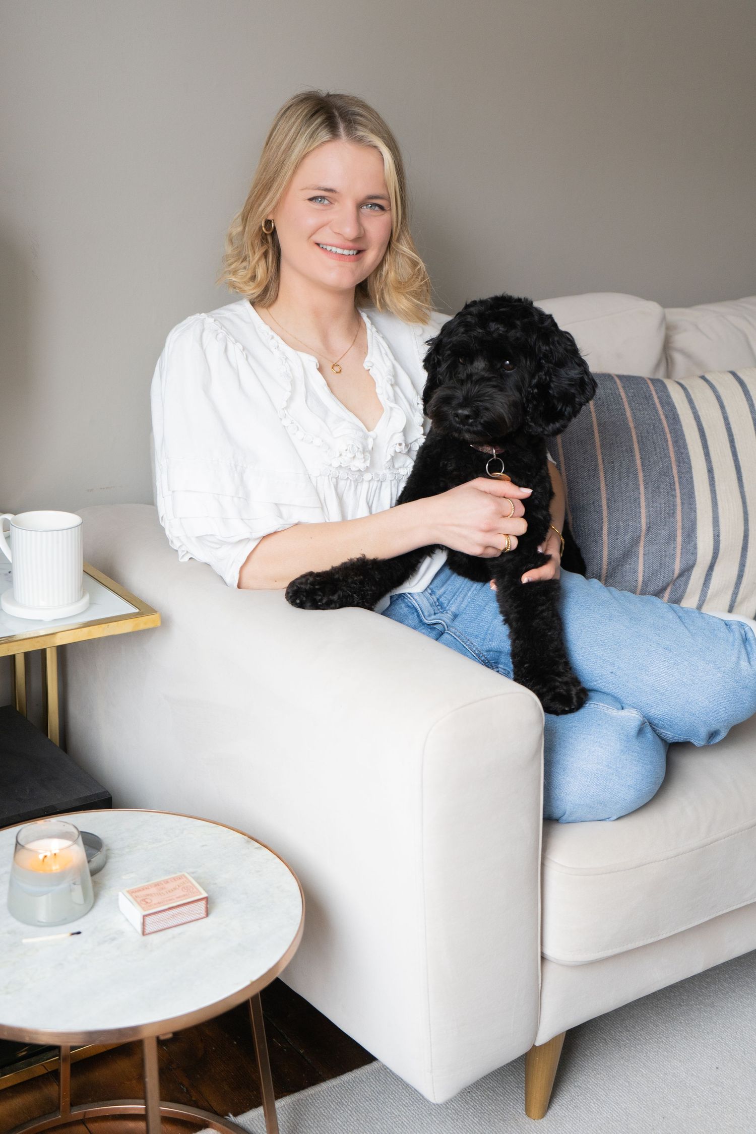 portrait of an artist sitting on her sofa dressed in white, smiling with her black dog sitting on her lap, neutral and light photography