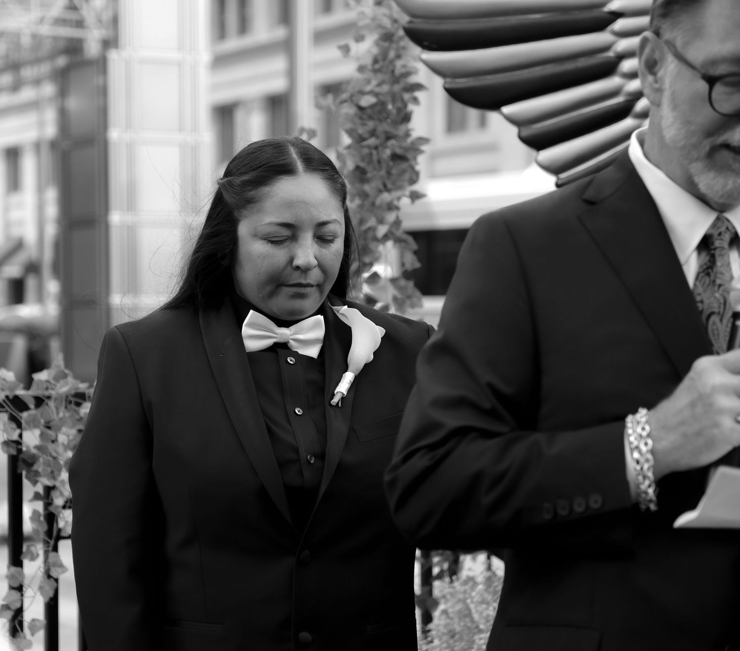 A black and white photograph shows figures in formal attire standing outside an establishment with a striped awning.