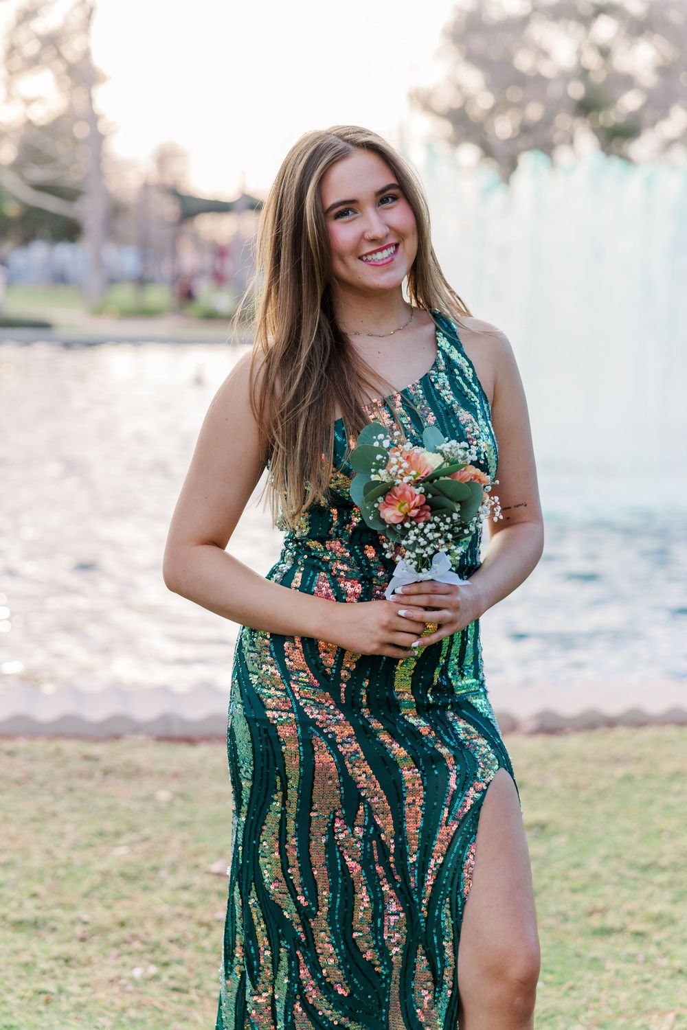 Someone in a green sequined dress with a thigh-high slit holds a bouquet of flowers at sunset by the beach.
