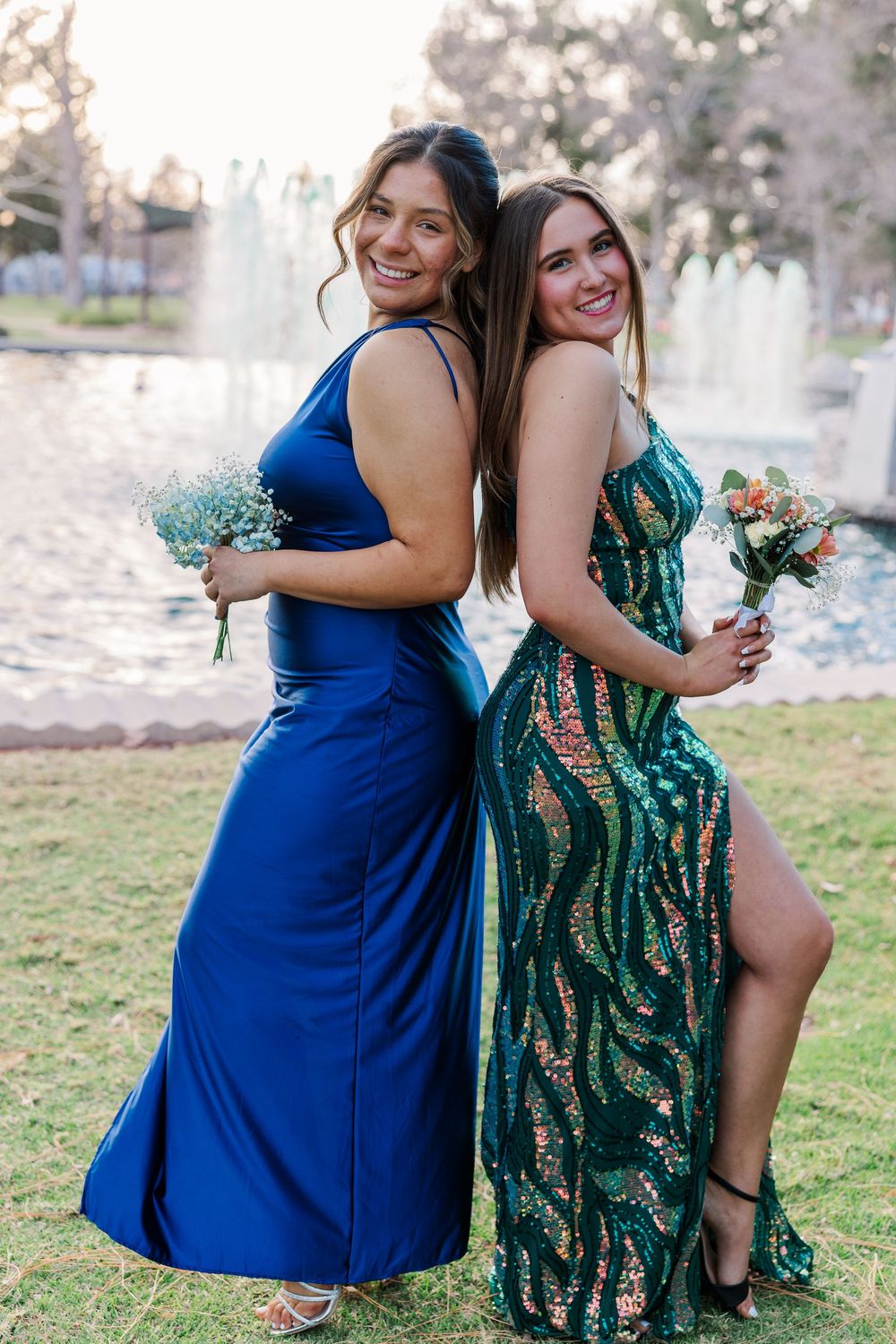 Two women in elegant evening gowns stand back-to-back holding floral bouquets in an outdoor garden setting at sunset.