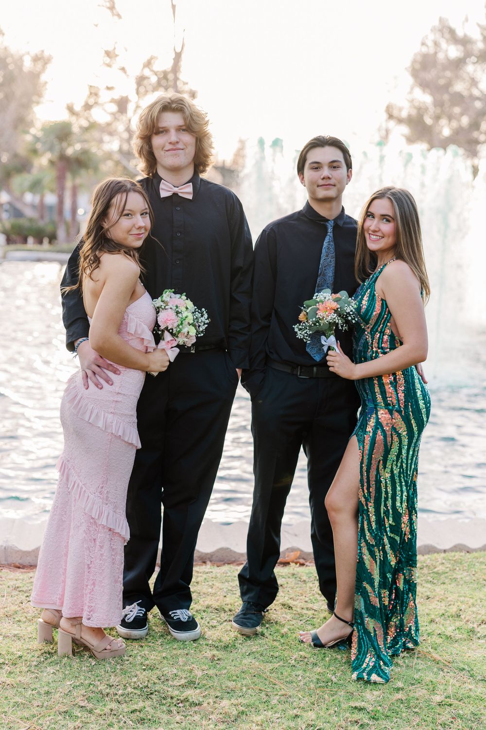 Prom couples in formal attire pose together outdoors with bouquets on a grassy area.
