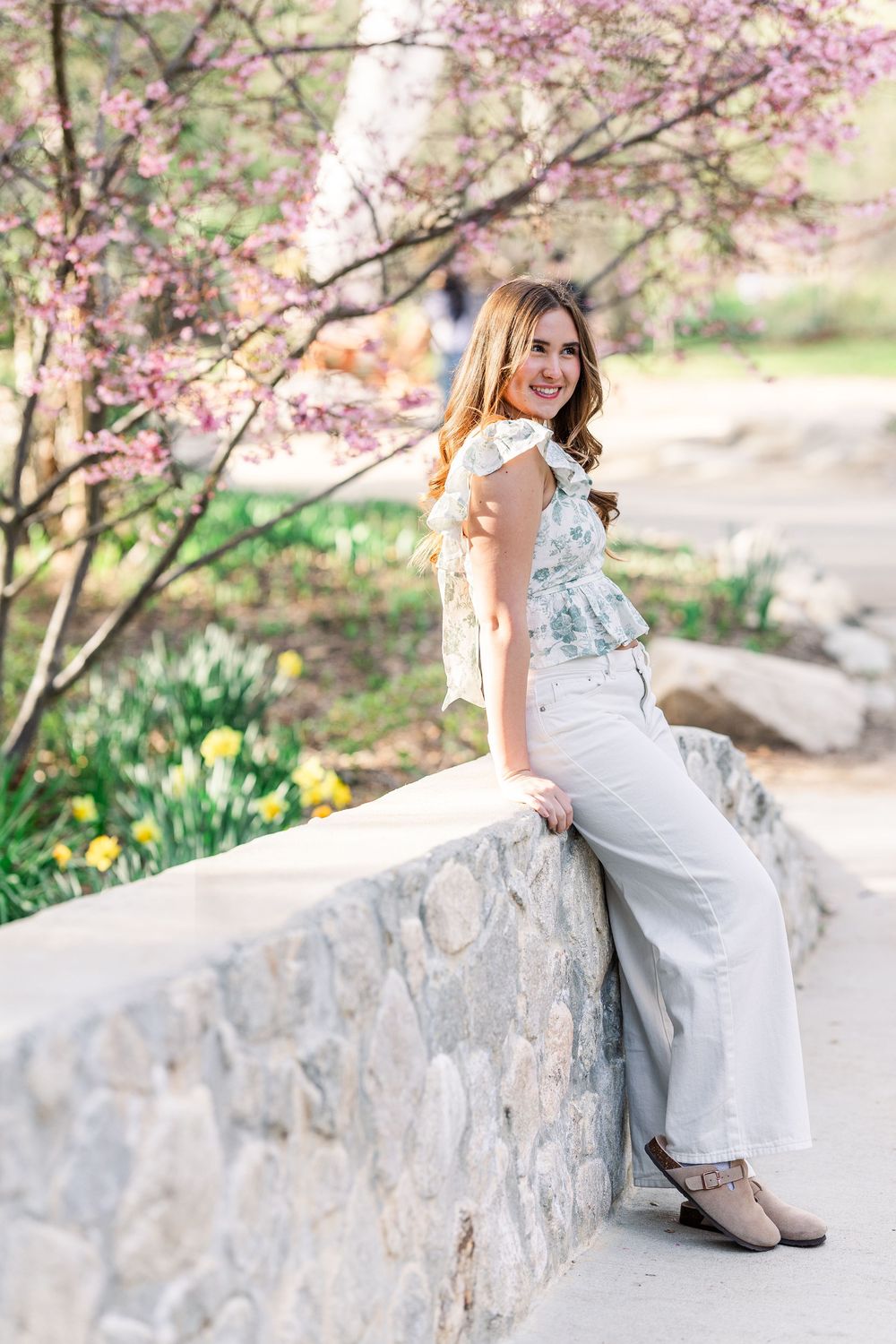 A figure in a white floral top and light pants sits on a stone wall beneath blooming pink cherry blossoms in spring.