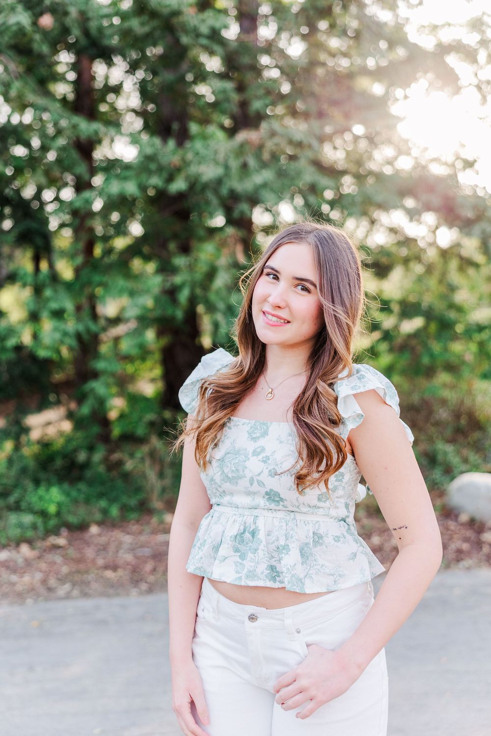 A person in a floral crop top and white pants poses outdoors with sunlit trees in the background.