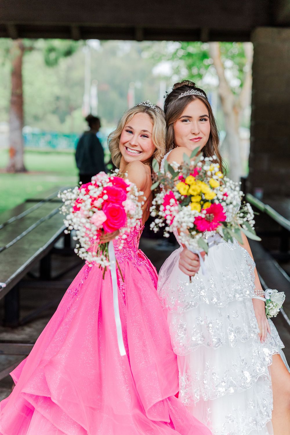 Bride in white lace dress and bridesmaid in vibrant pink gown holding colorful bouquets under gazebo.
