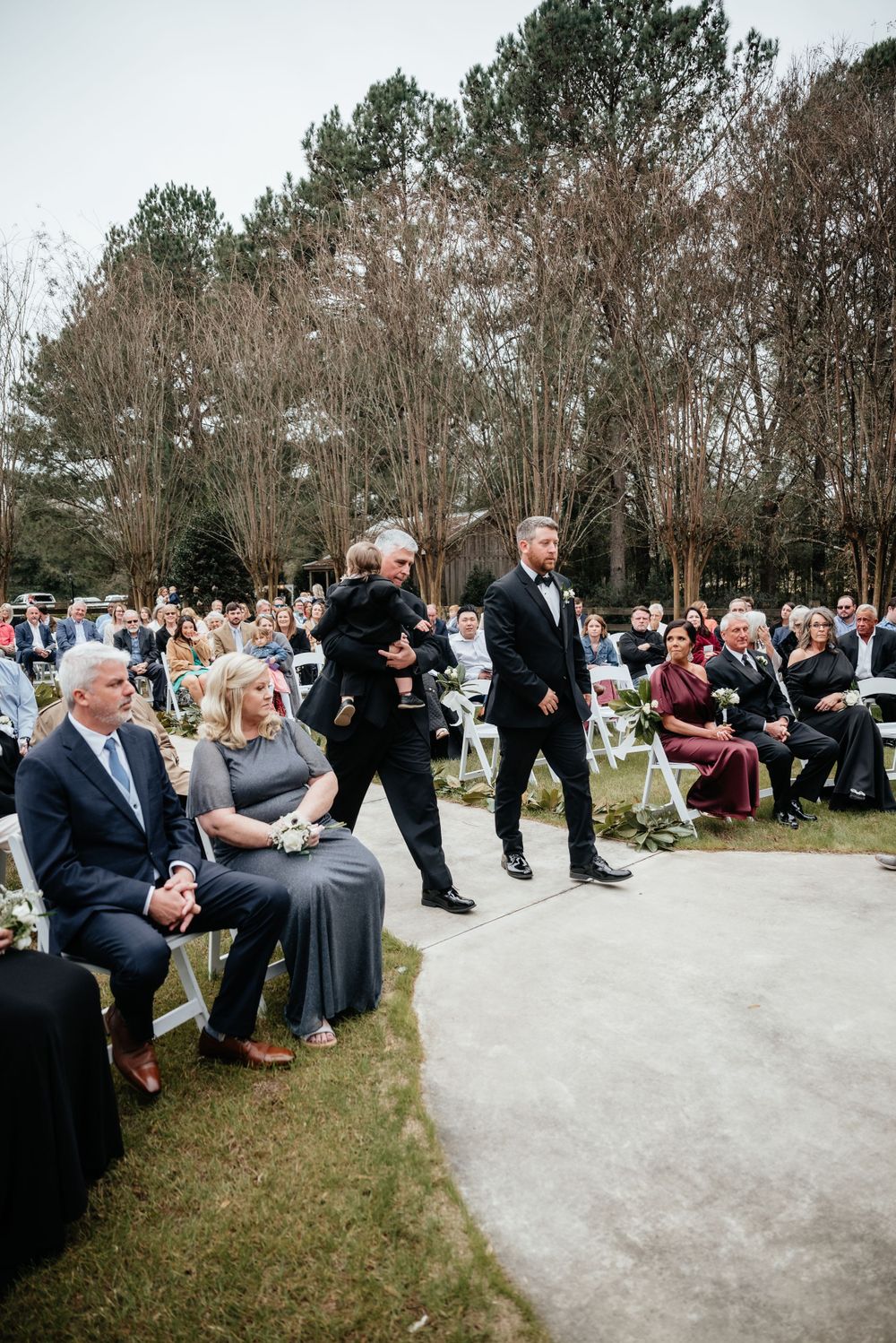 Groom walks down aisle with child at elegant outdoor wedding ceremony with seated guests.