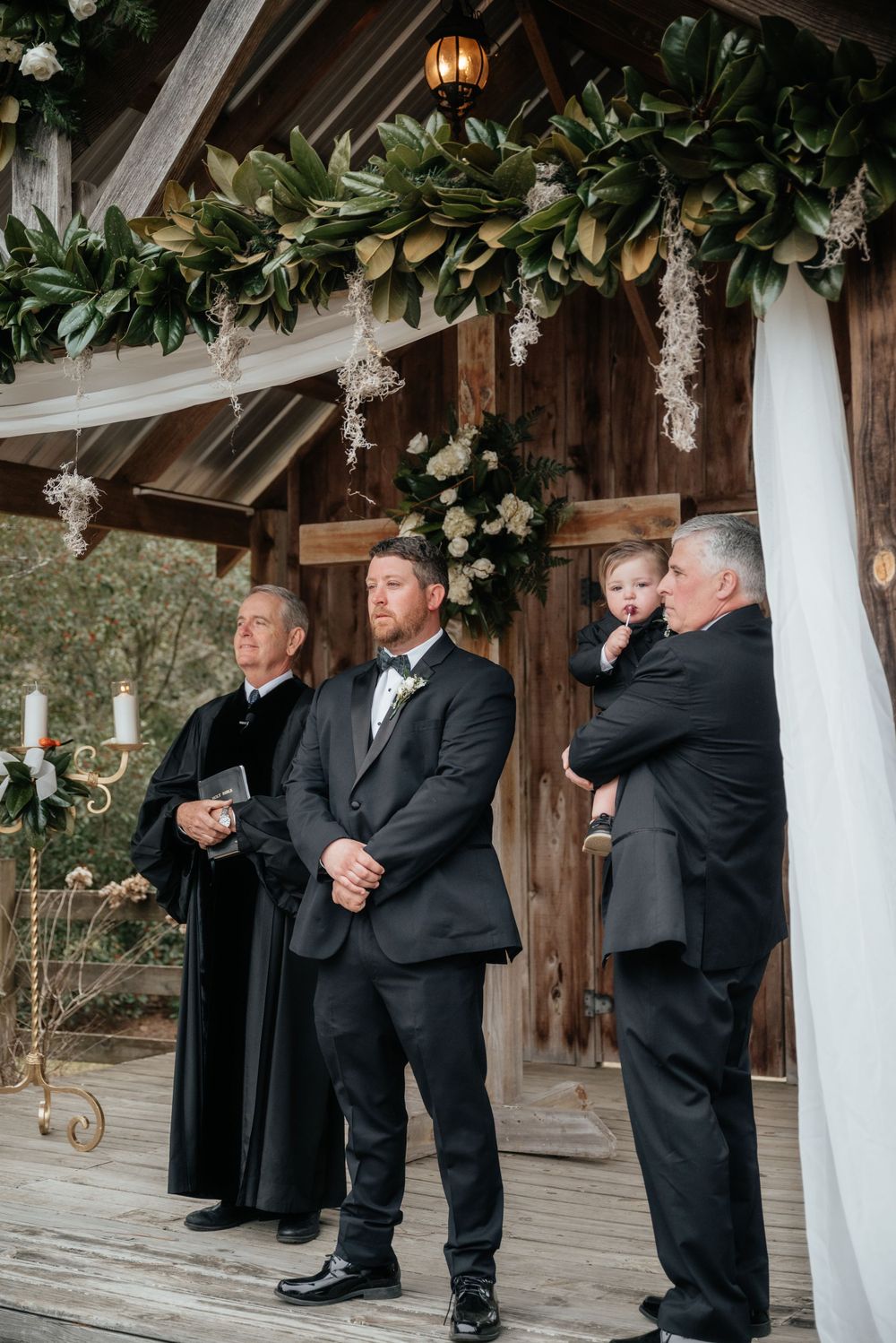 Groom stands at altar under floral arch with officiant and groomsmen at outdoor barn wedding.