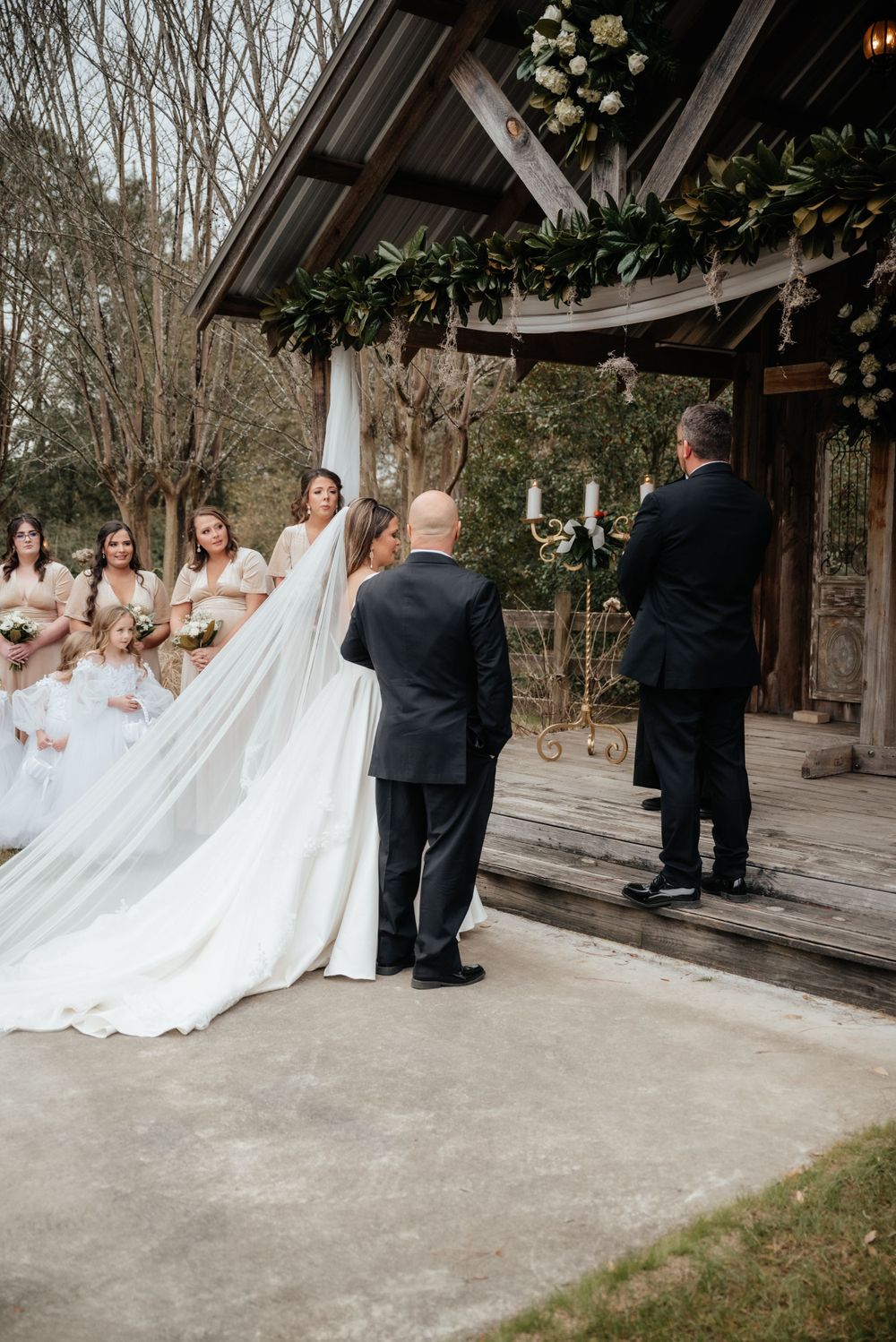 Bride in flowing white gown with long veil approaches groom at rustic outdoor wedding ceremony.