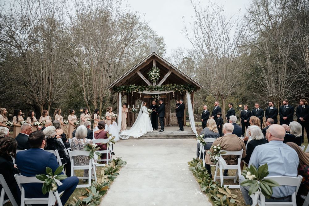 Outdoor wedding ceremony at rustic wooden chapel with guests seated along greenery-lined aisle.