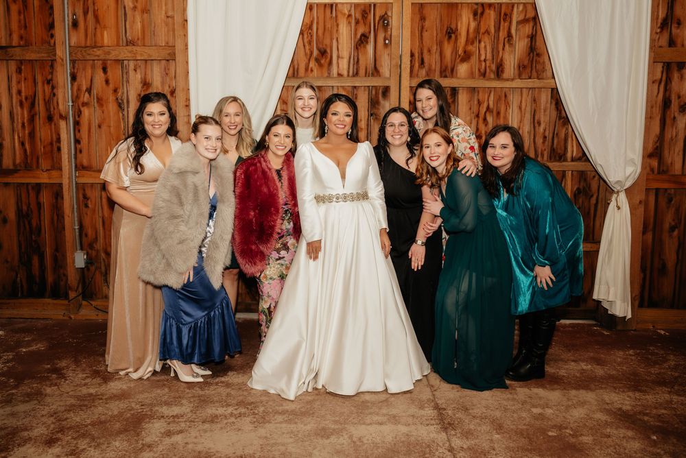 Bride in elegant white gown poses with bridesmaids and guests inside rustic barn wedding venue.