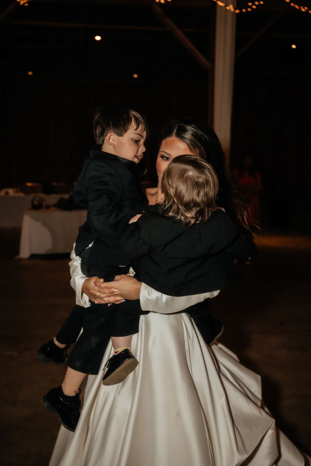 Bride in white satin gown lovingly holds two young boys in black suits at wedding reception.