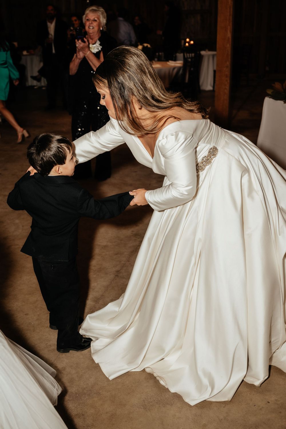 Bride in white satin gown dances joyfully with young boy in black suit at wedding reception.
