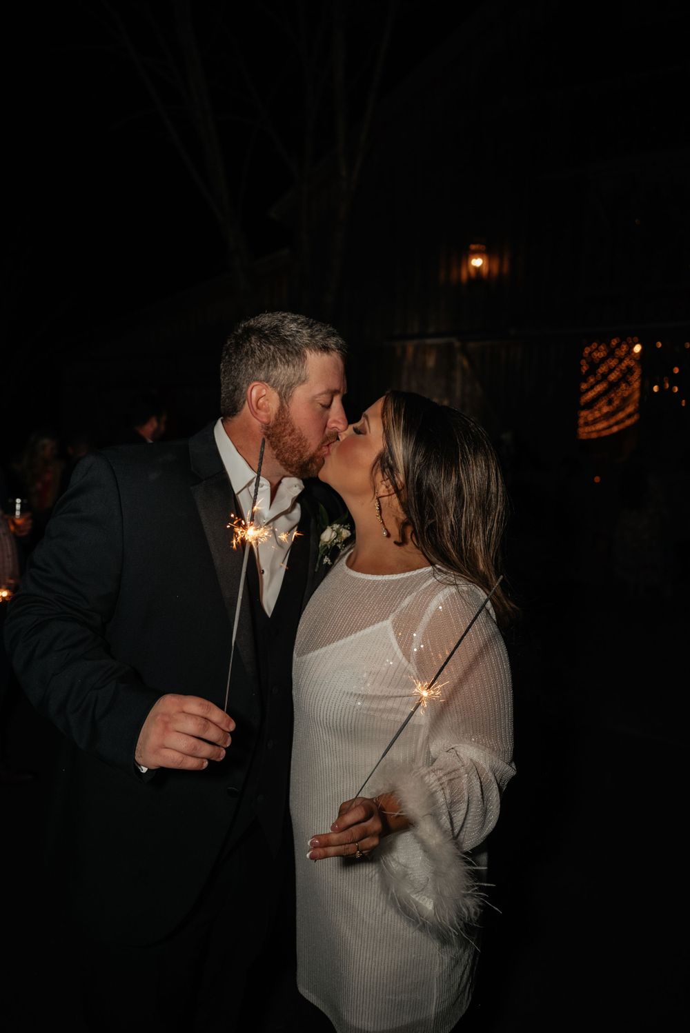 Newlyweds share romantic kiss while holding sparklers during nighttime wedding send-off celebration.