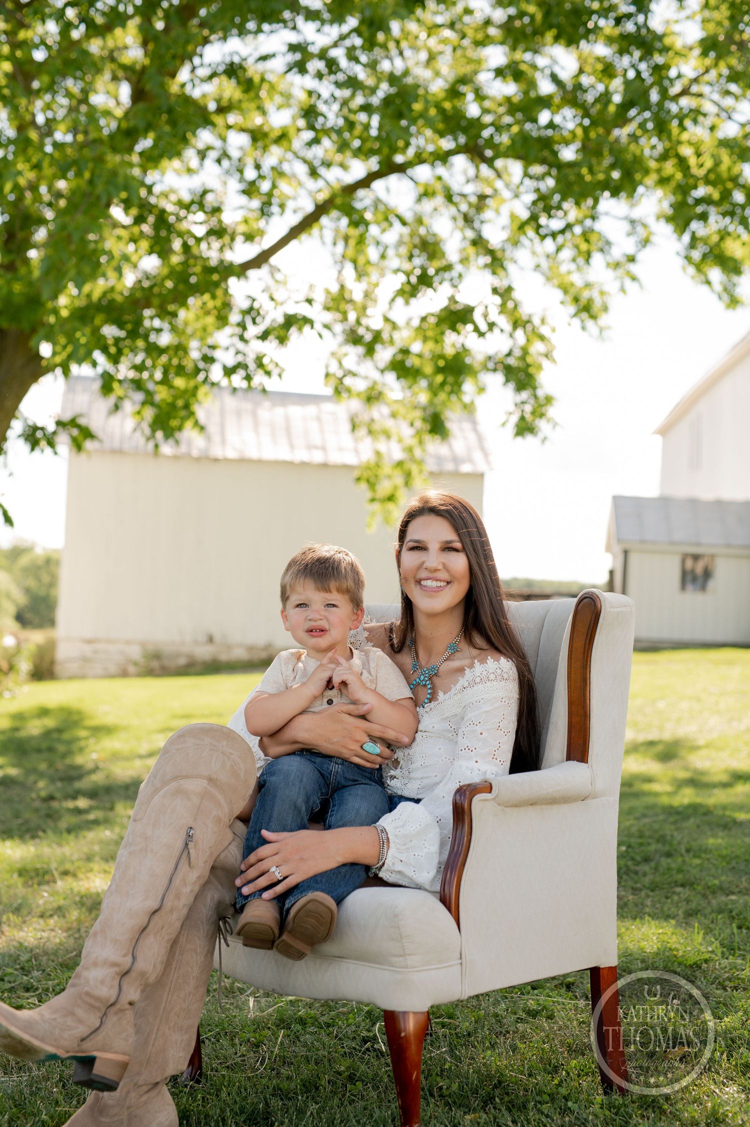 An outdoor family portrait features a duo sitting in a vintage white chair against white barns and green trees.