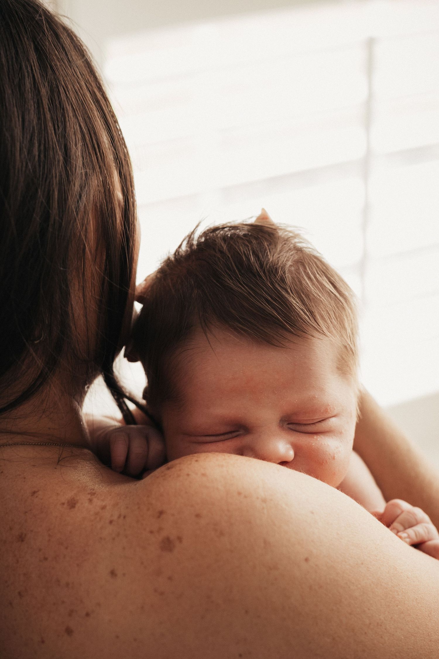 Tender moment between parent and sleeping newborn baby resting on bare shoulder in soft window light.